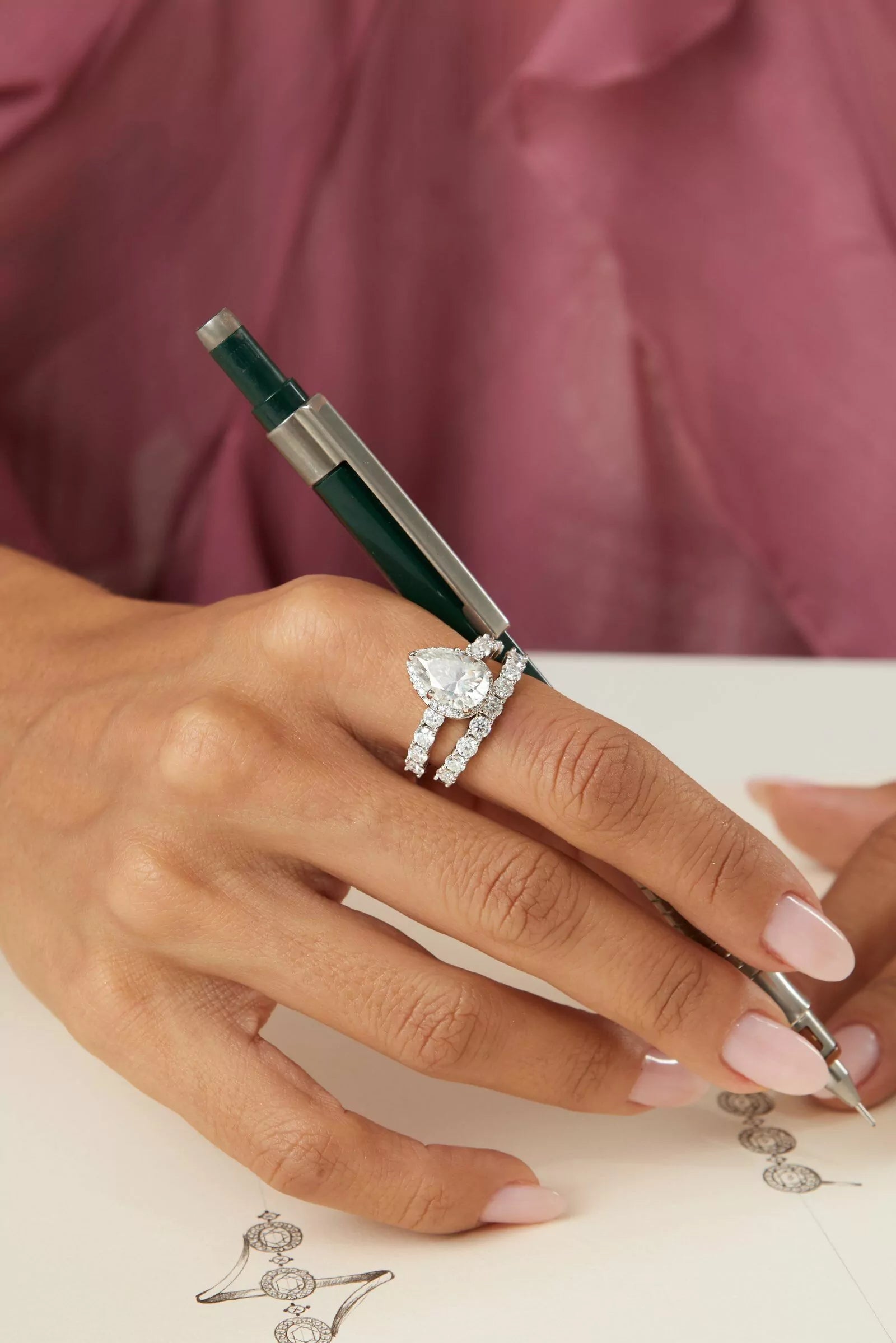 Hand wearing a diamond ring with a pen on a pink fabric background